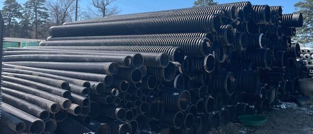 Stacked black plastic pipes stored outdoors in a yard, with trees and blue sky in the background