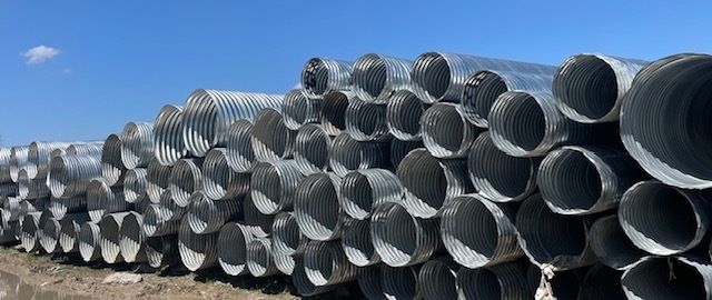 Stacks of large gray corrugated metal pipes lined up outdoors under a blue sky