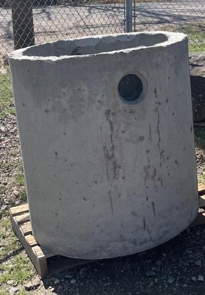 Large concrete cylindrical tank with a round black opening, sitting outdoors on pallets near a fence.