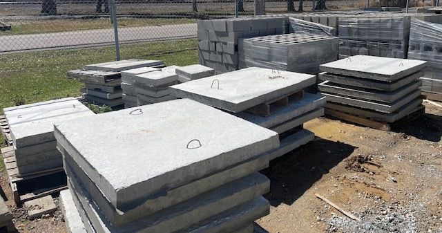Stacks of gray concrete slabs on pallets in a dirt lot near a road