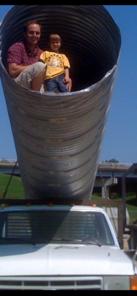 Two people sitting inside a giant metal pipe mounted on a white truck outdoors