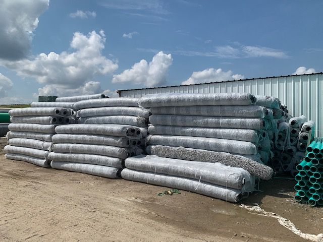 Stacks of wrapped drainage pipes and green pipe coils beside a metal building under a cloudy sky