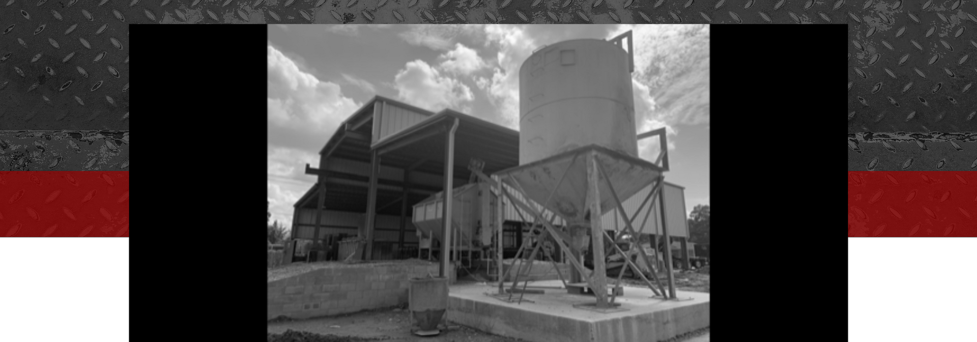 Grayscale industrial facility with metal framework, large tank, and silo under a cloudy sky