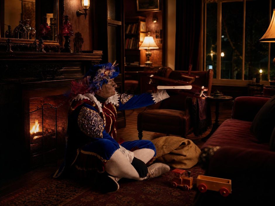 Een blauwe knuffelwolf zit bij de open haard in een gezellige, schemerig verlichte woonkamer met een brandende lamp 