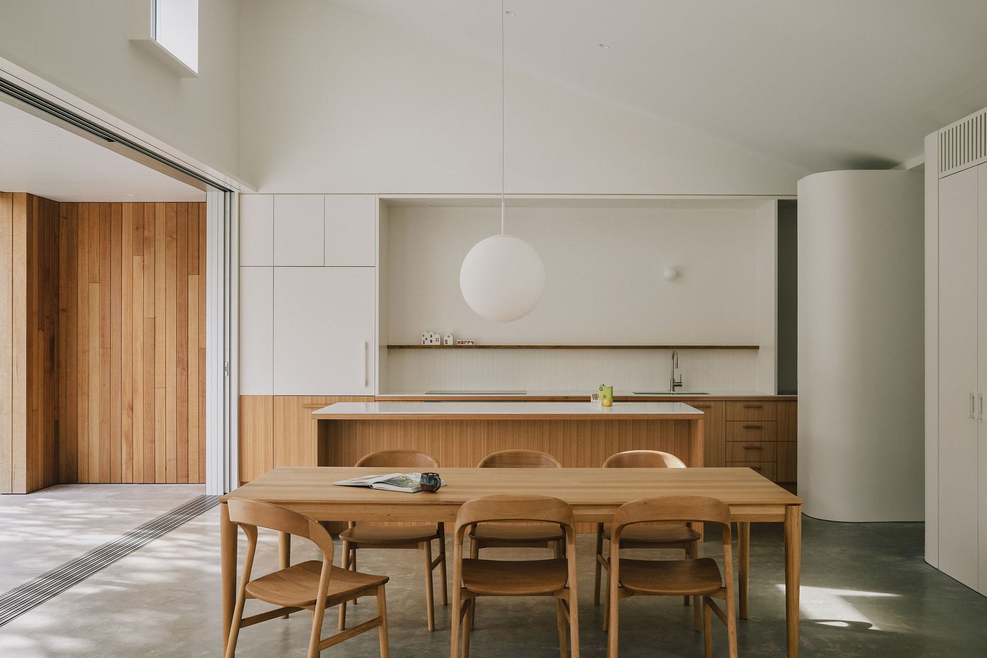 Modern minimalist kitchen and dining area with wooden table, chairs, white walls, and a hanging globe light