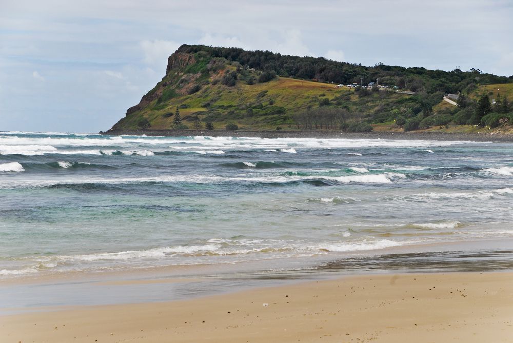 A Beach With A Mountain In The Background And Waves Crashing On The Shore — Premier Roofing Solutions In Lennox Head, NSW