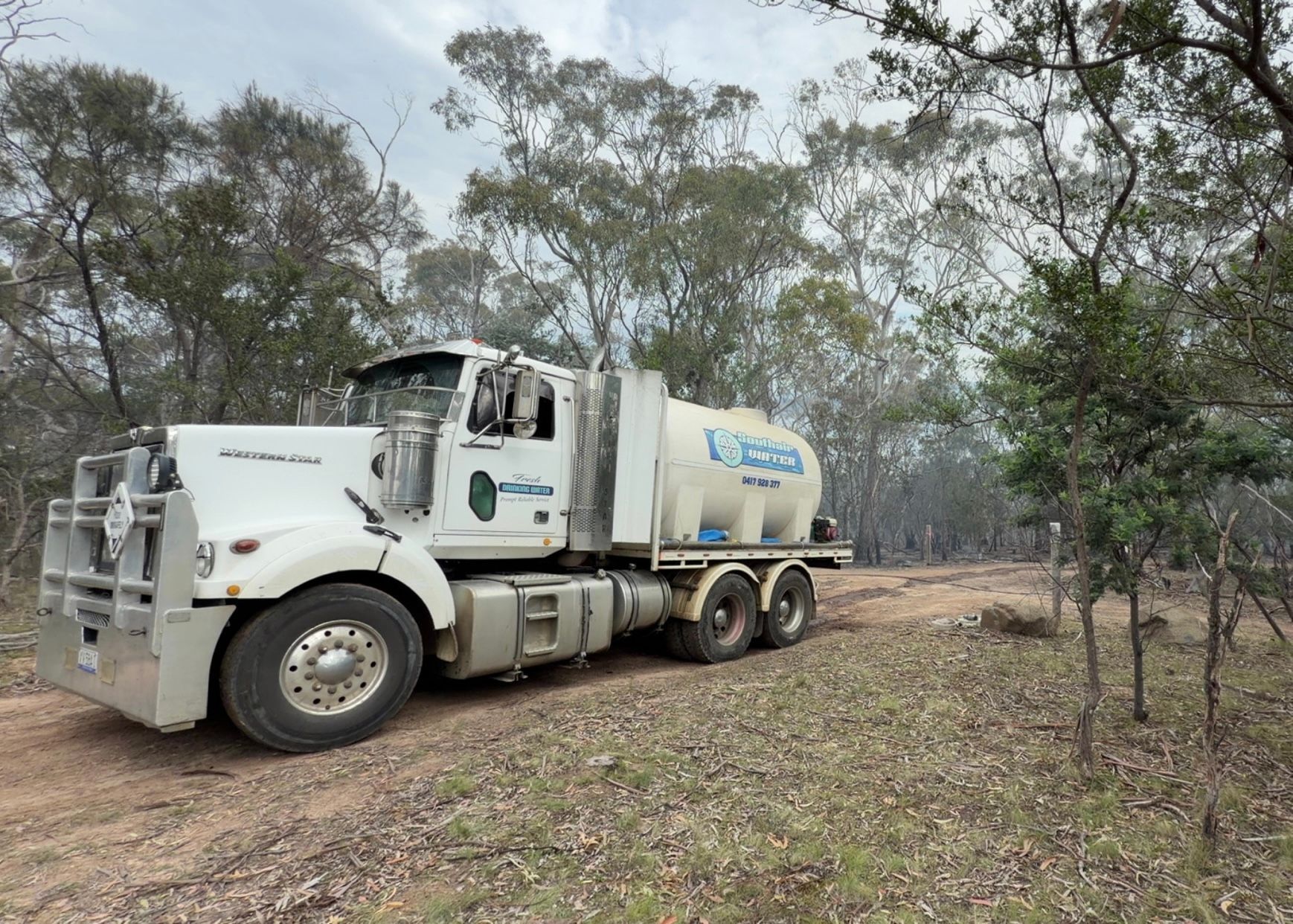 White tanker truck driving on a dirt path through sparse trees and brush