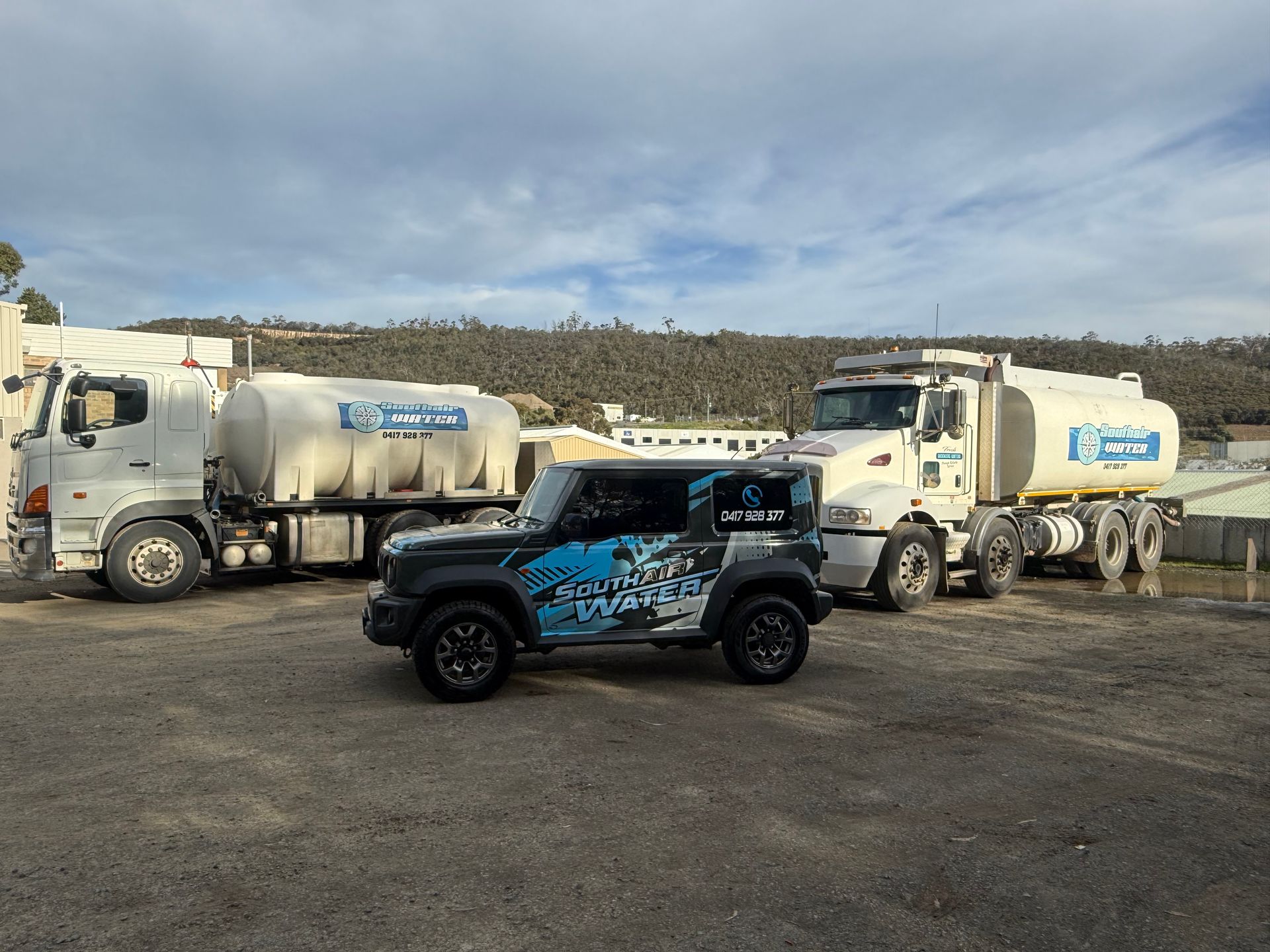 Black off-road Jeep in front of two white tanker trucks in a dusty lot under a cloudy sky