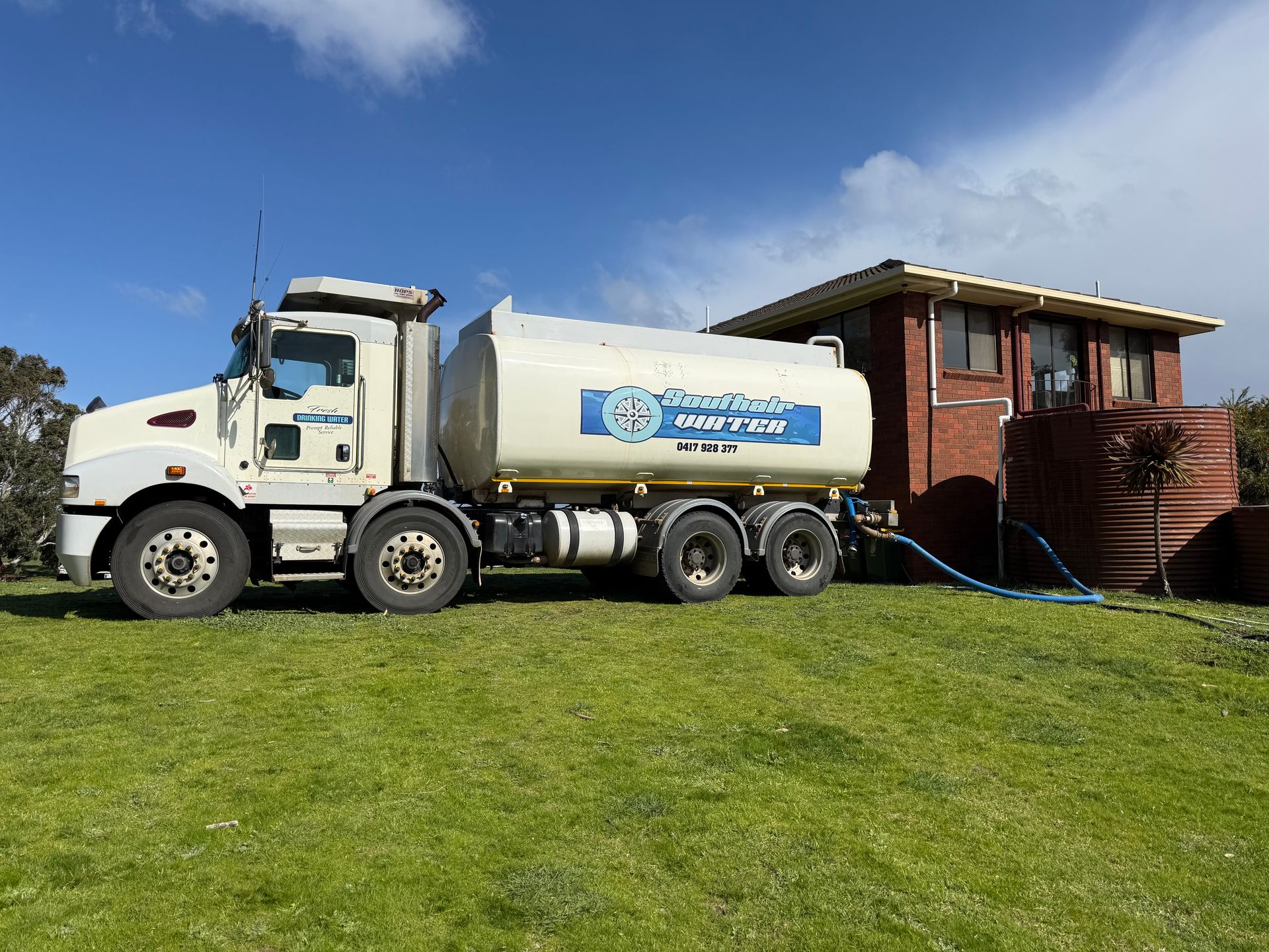 White tanker truck beside a rustic wooden building on a grassy hill under a blue sky