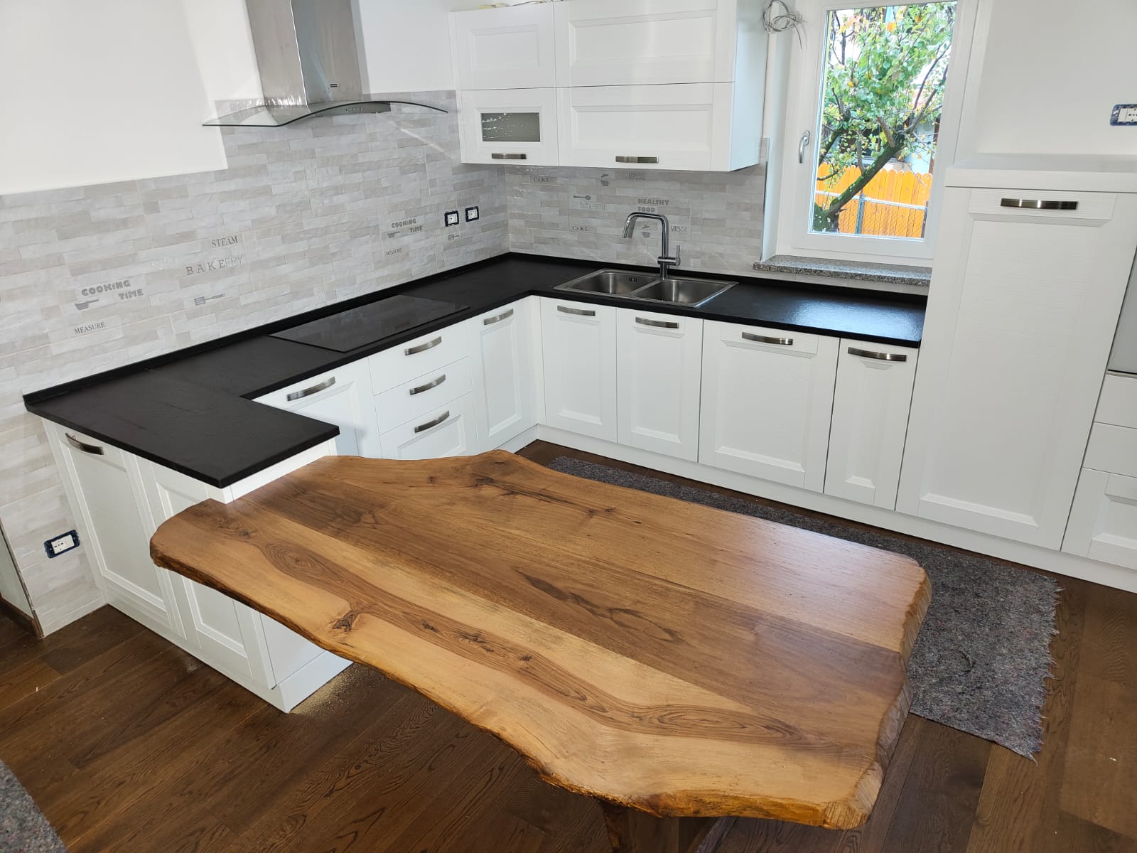 Cucina moderna con mobili bianchi, ripiani neri, paraschizzi grigio e un'isola con tavolo in legno.