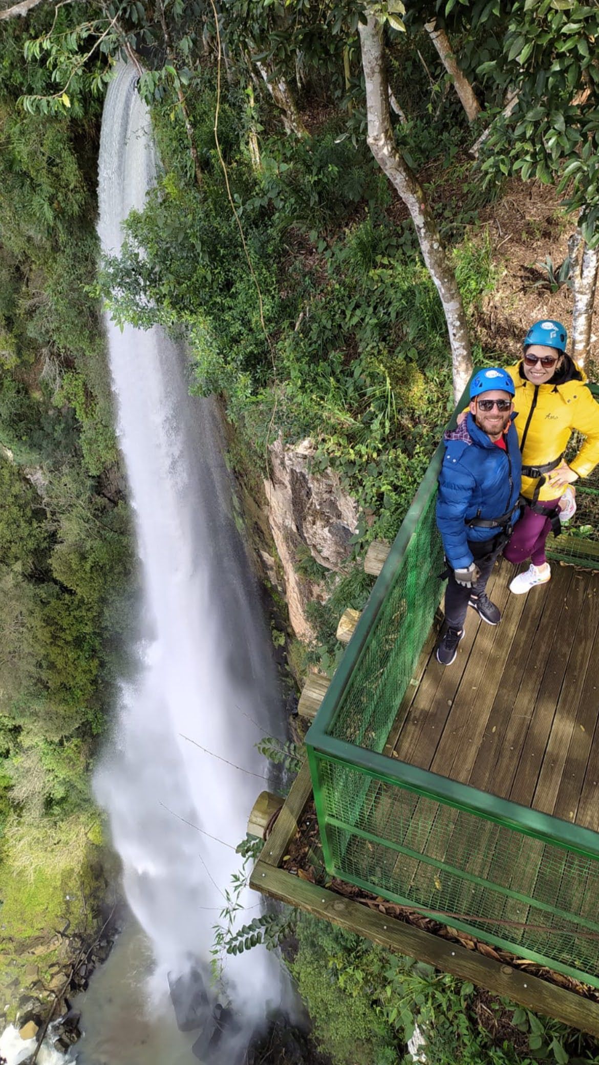 Duas pessoas em uma plataforma perto da cachoeira. Uma com jaqueta azul, a outra com jaqueta amarela. Folhagem verde, corrimão de metal.