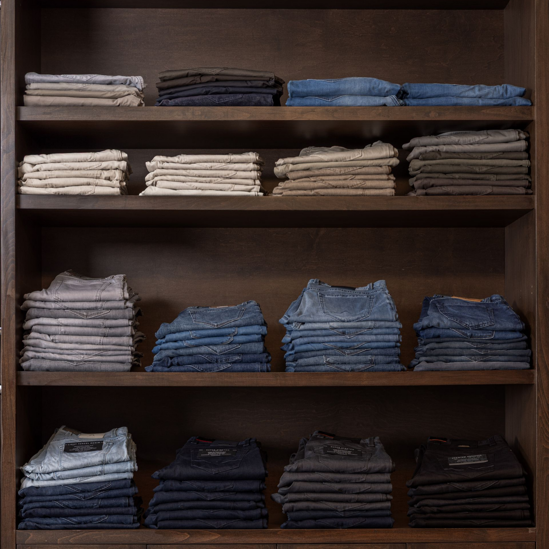 Shelves displaying folded jeans in various shades of blue, gray, and brown. Wooden cabinet.