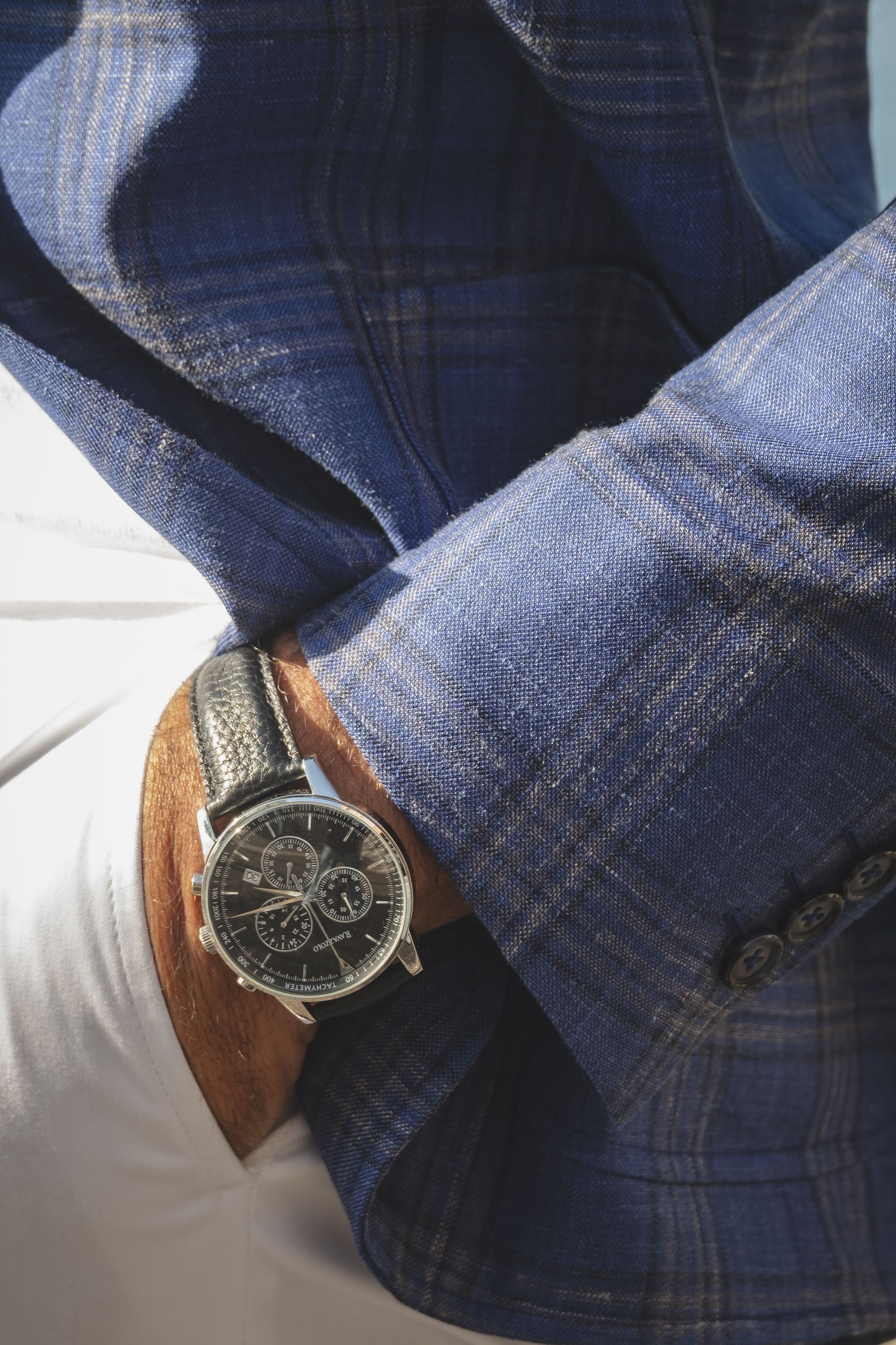 Man's wrist with a luxury watch peeking out of a blue plaid blazer sleeve. White pants and sunlight are visible.