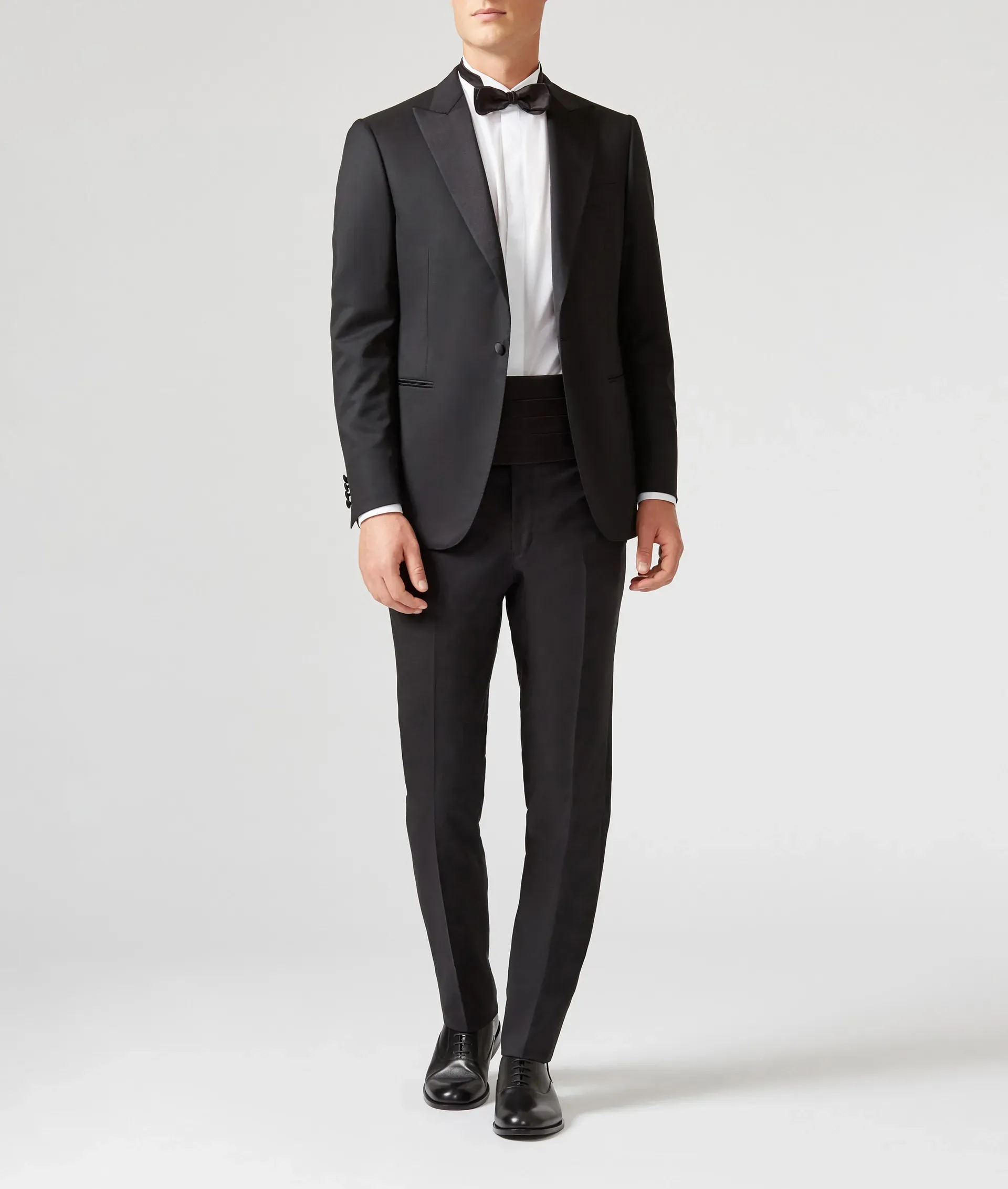 Man in black tuxedo with bow tie and cummerbund, standing against a neutral background.