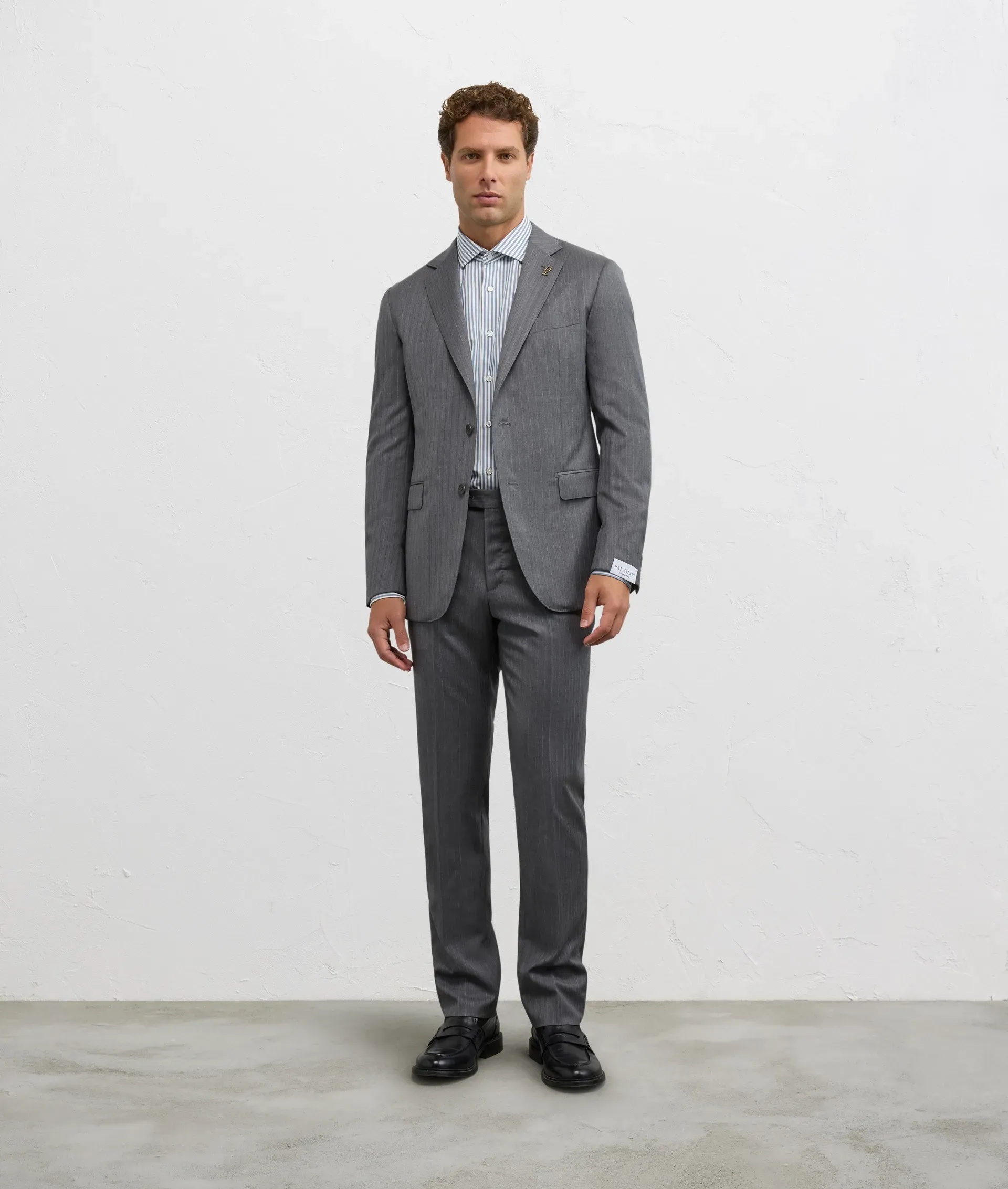 Man in a gray suit and striped shirt stands in front of a white wall.