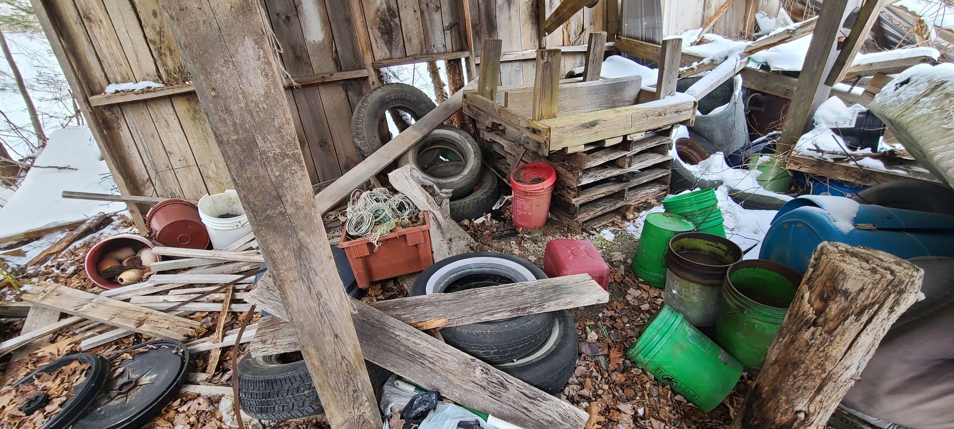 Cluttered outdoor shed with tires, buckets, tools, and green plastic containers among wooden debris