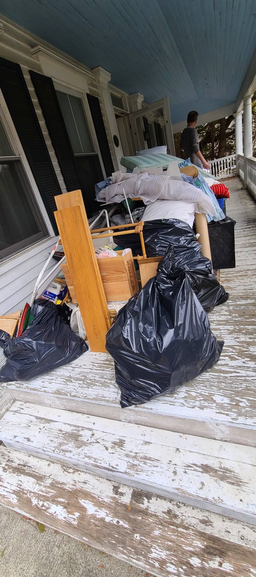 Porch piled with trash bags, bags, and debris beside a house with weathered flooring.