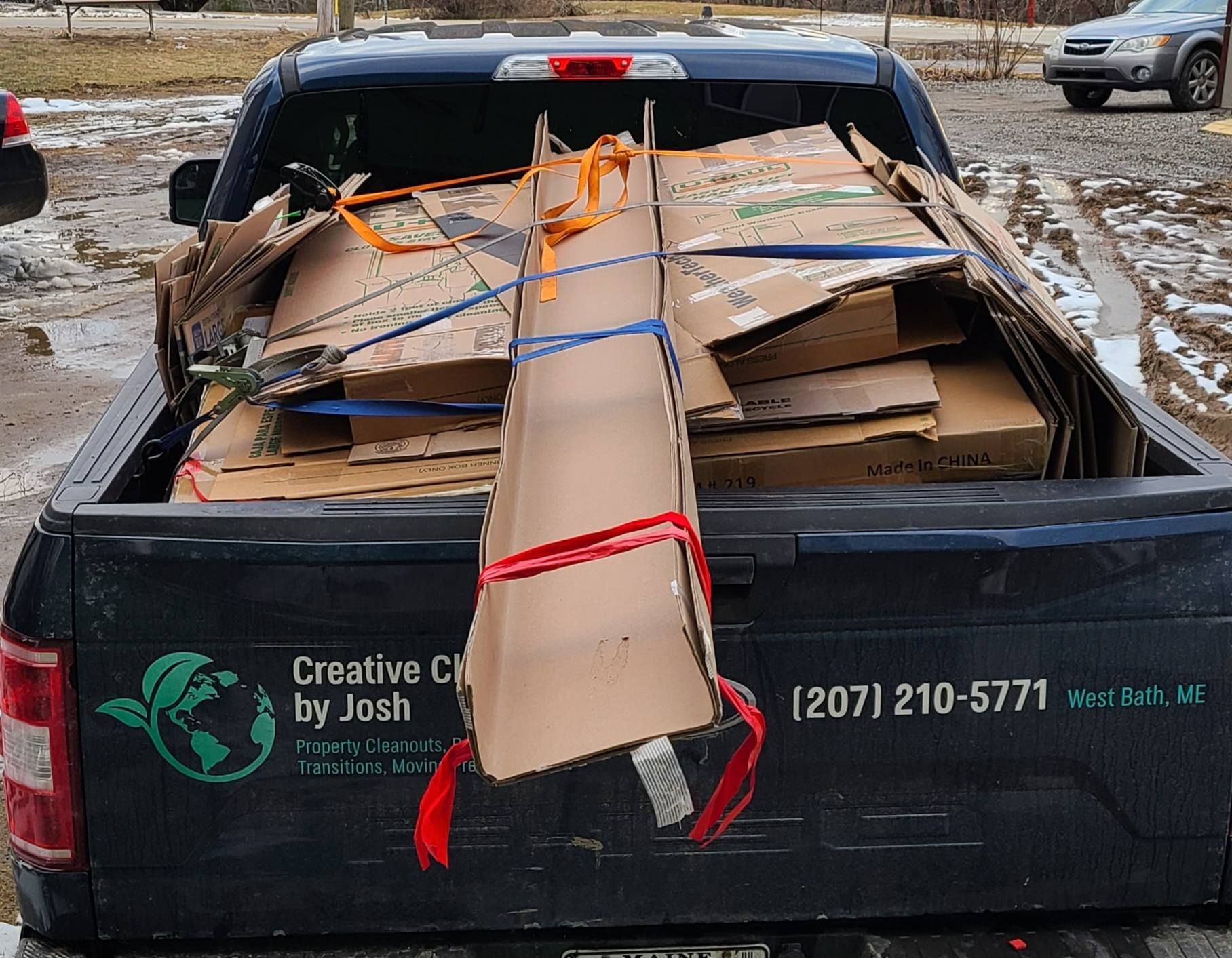 Pickup truck bed loaded with tied-down cardboard and scrap on a muddy roadside