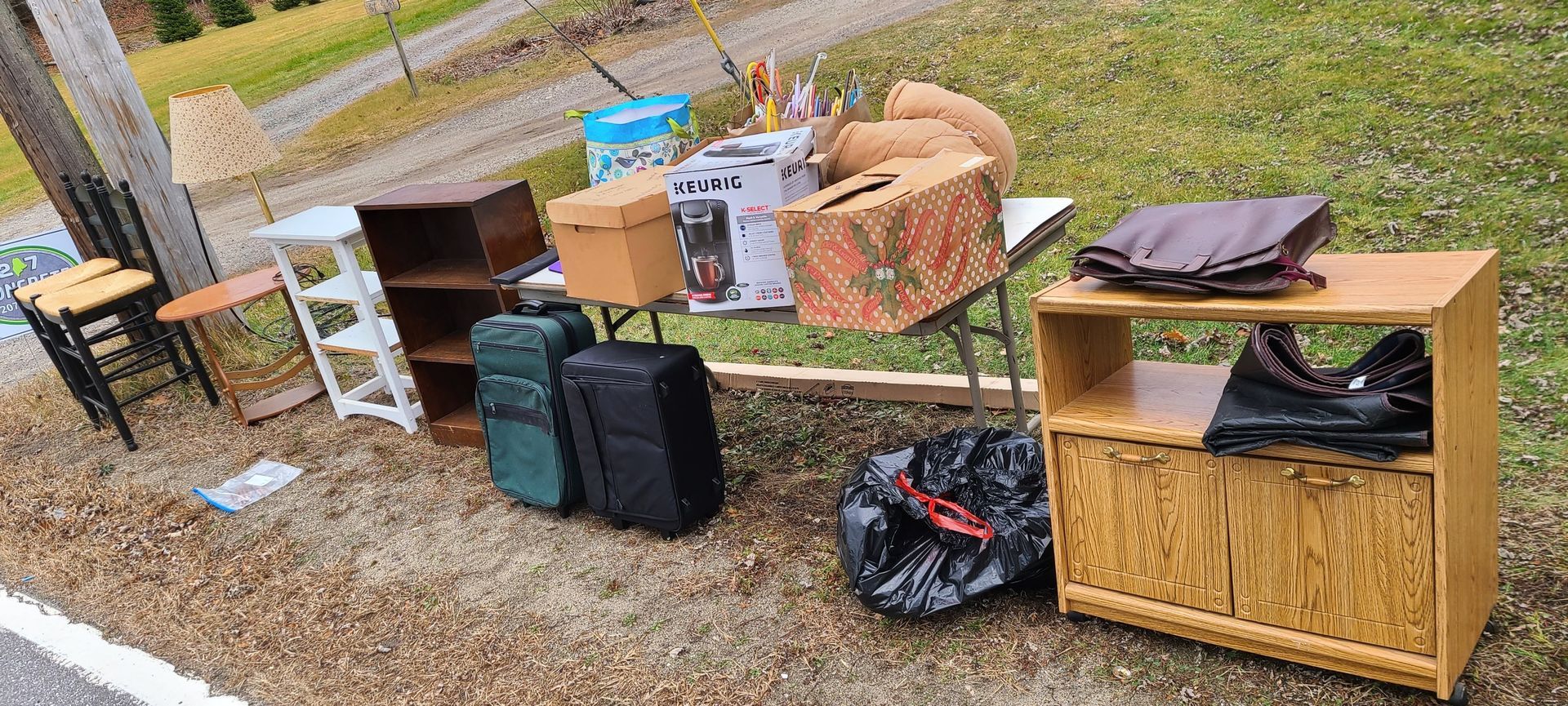 Outdoor yard sale setup with tables of assorted items, boxes, bags, and a wicker cabinet on gravel.