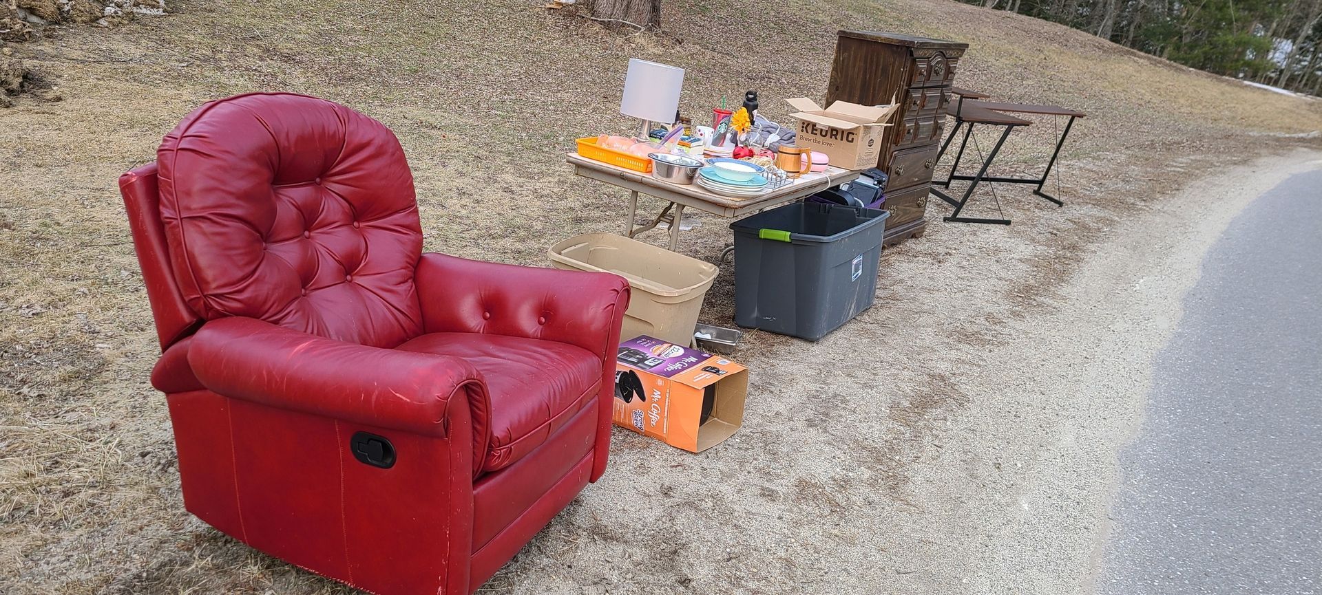 Red recliner chair beside a roadside table with assorted items on gravel.