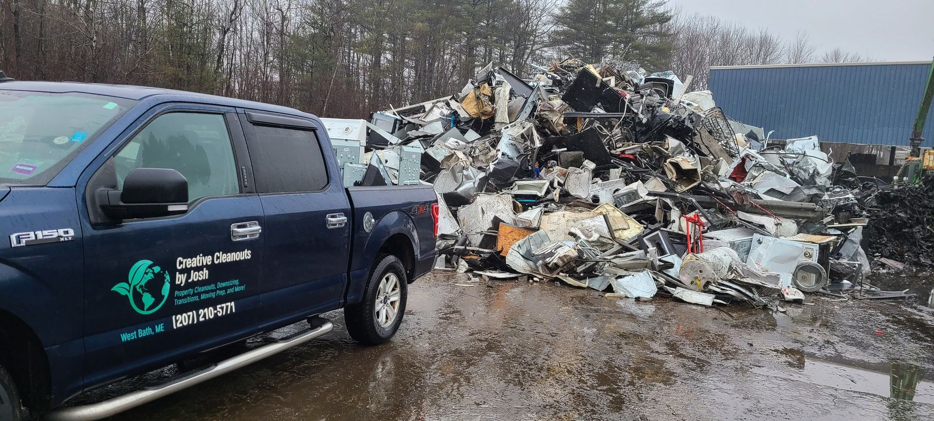 Dark blue pickup truck beside a large pile of debris and trash at a muddy roadside.