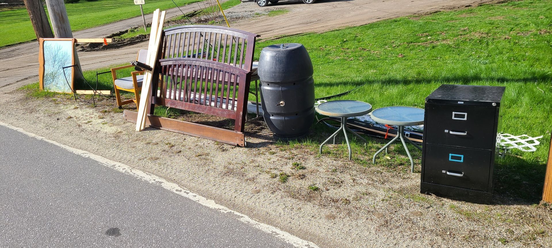 A roadside collection of assorted trash bins, a chair, a barrel, and a folding cot on a grassy verge