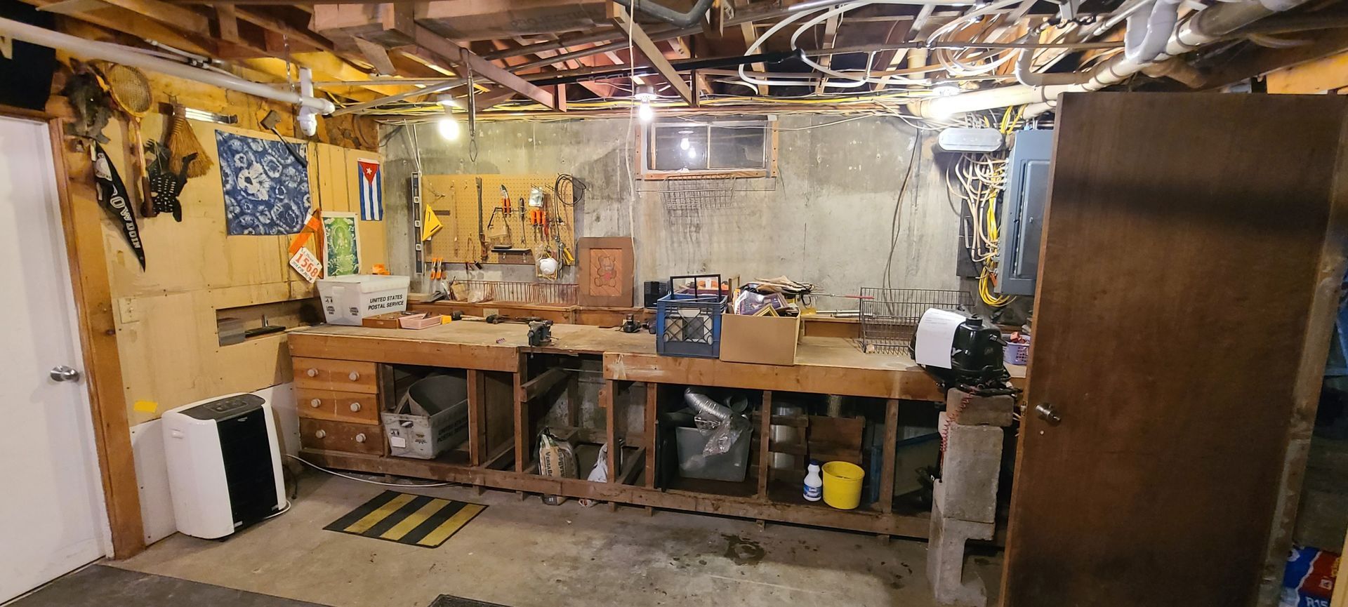 Cluttered basement workshop with workbench, tools, exposed ceiling, and storage shelves.