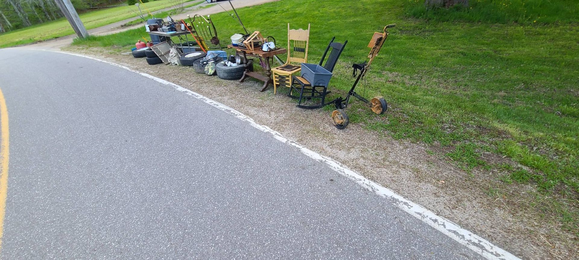 Lawn chairs and yard sale items set up beside a curving road on a grassy roadside.