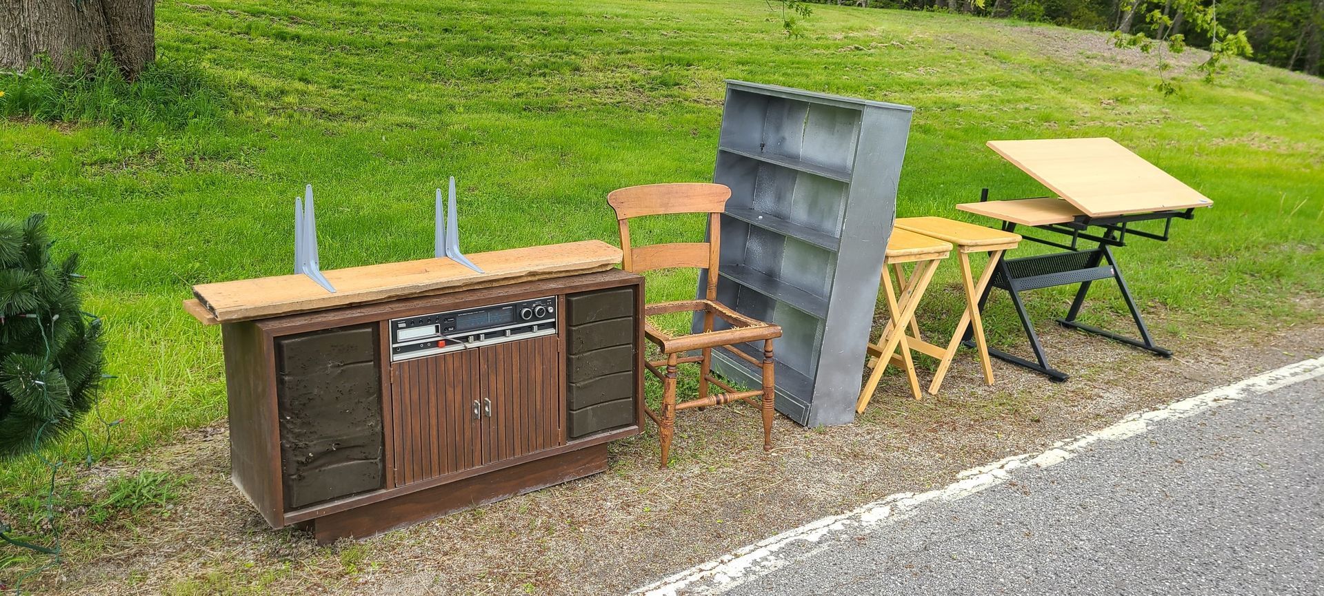 Outdoor roadside setup with a wooden cabinet, chair, folding table, and metal panel on a grassy verge