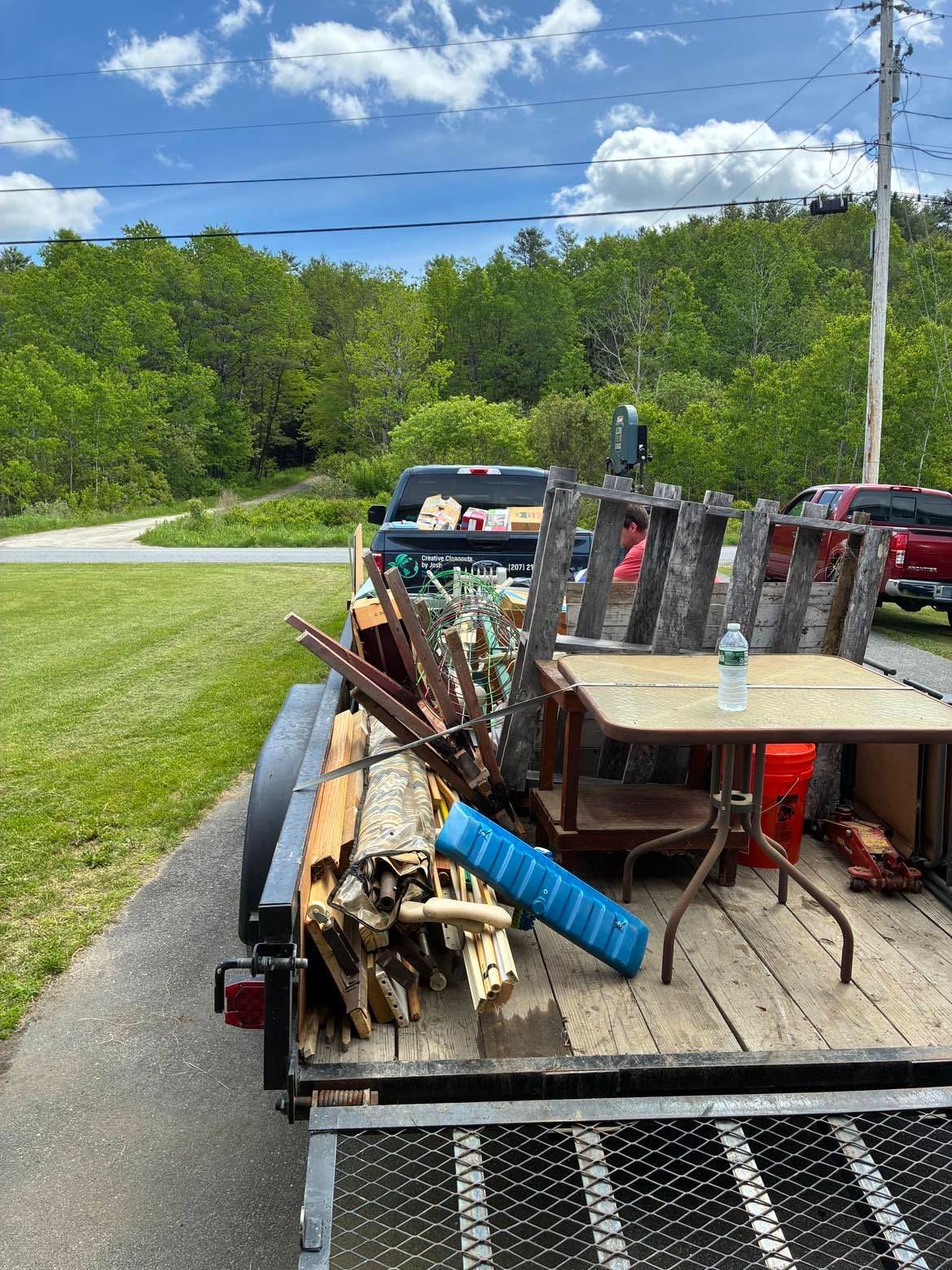 Loaded utility trailer with scrap wood and metal, parked on a roadside by green trees and a blue sky