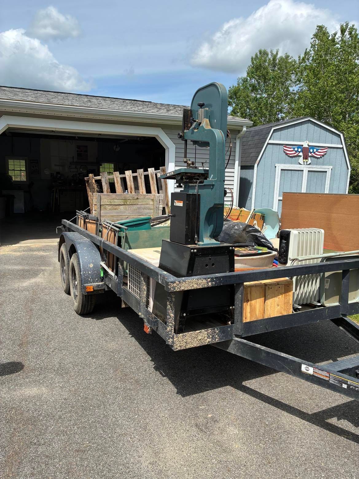 Black trailer loaded with a pallet jack and tools in front of an open garage and shed