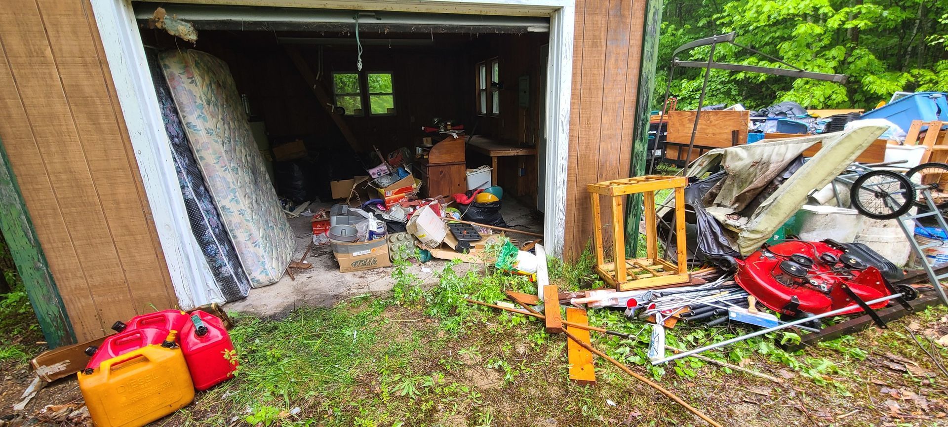 Cluttered garage doorway with assorted tools, bins, and scrap piled outside on a grassy, overgrown area