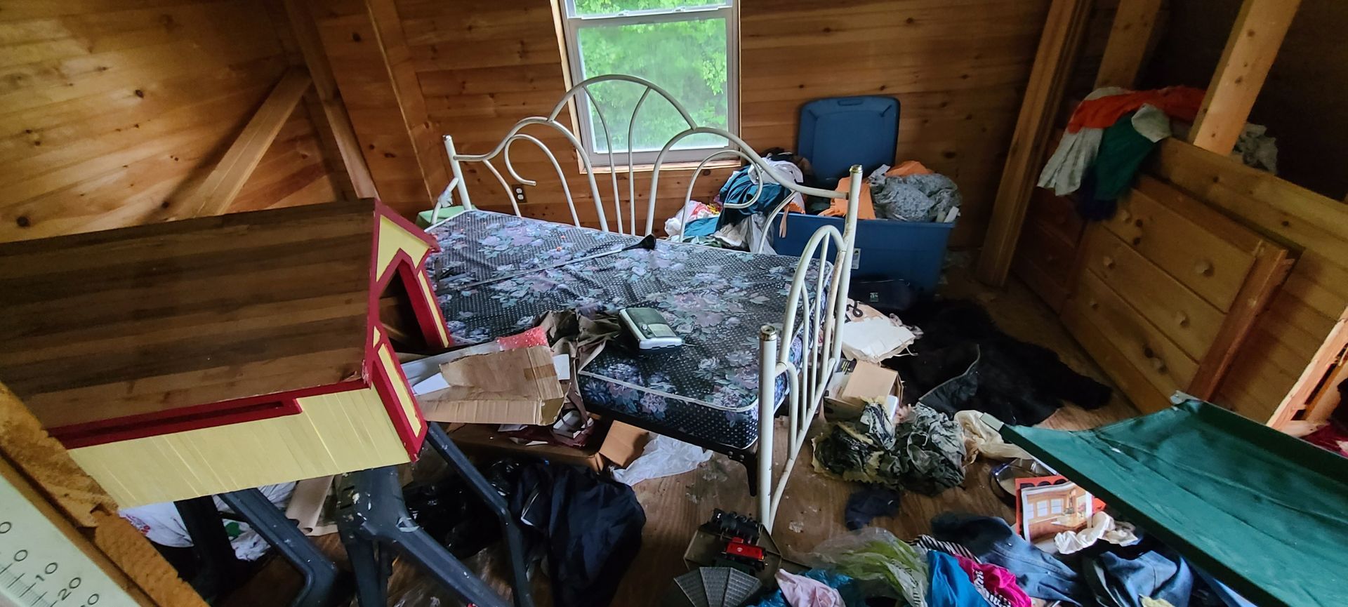 Messy attic bedroom with a bed, scattered clothes, and boxes under a small window