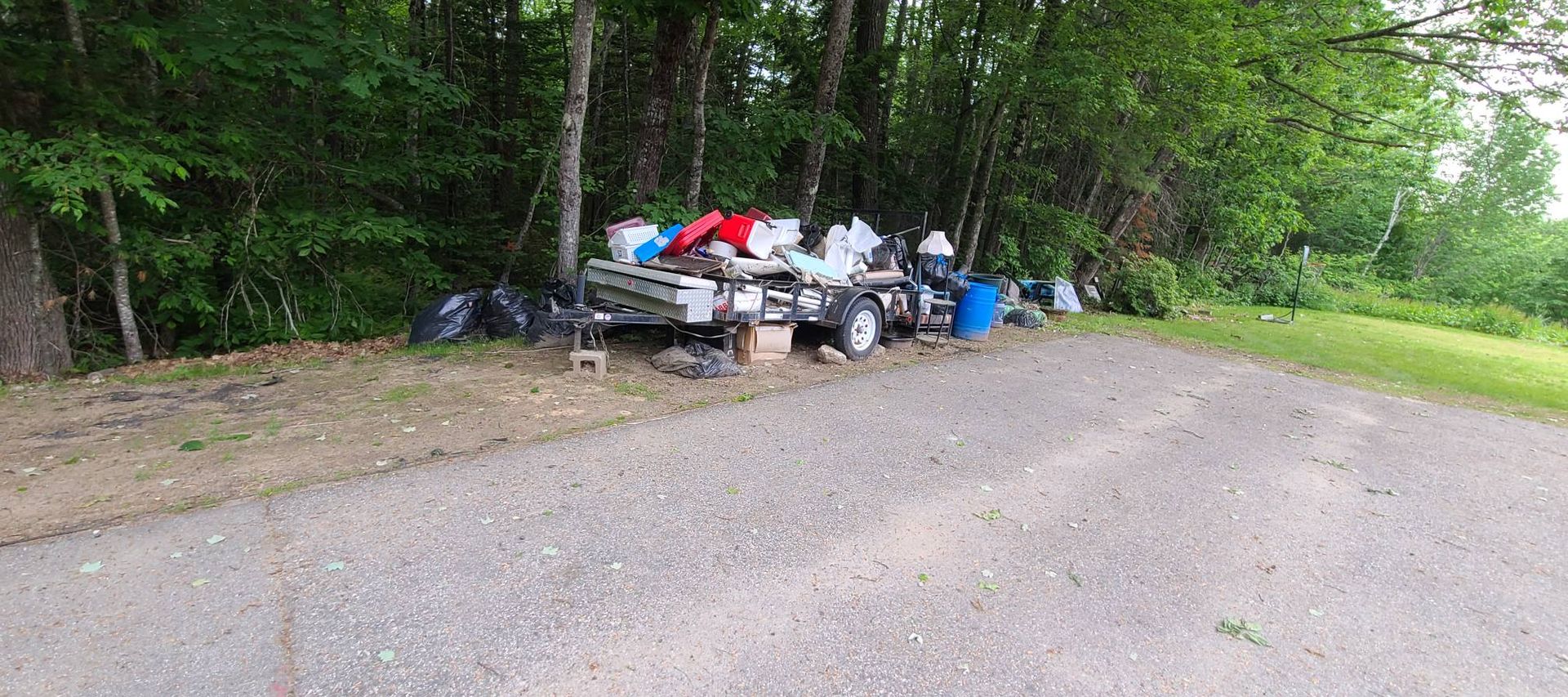 Overflowing trash and debris piled beside a road at the edge of a wooded area.