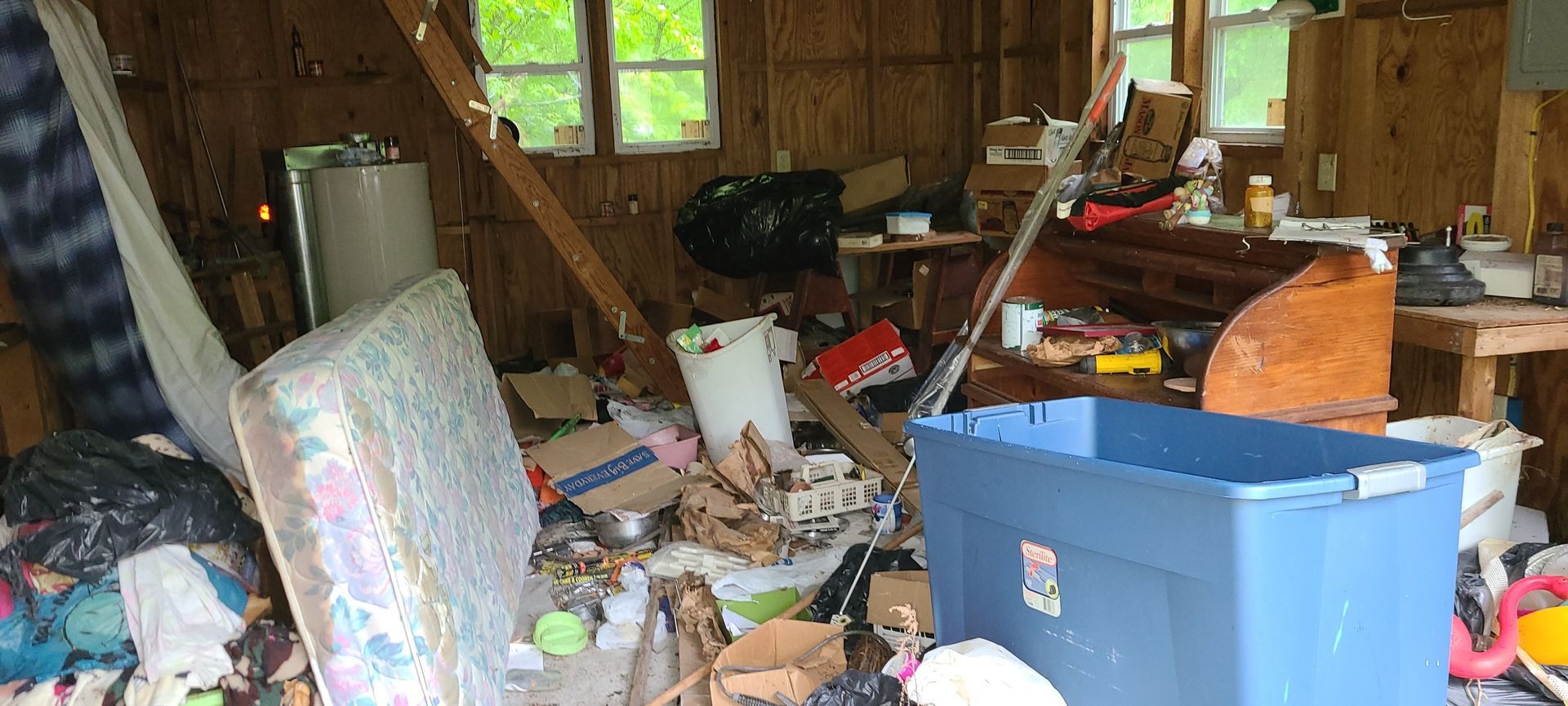 Cluttered, messy room with scattered trash, boxes, and a large blue bin under a window.