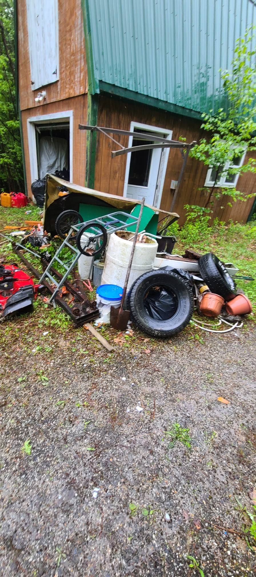 Cluttered yard by a tilted green-and-brown shed, with tires, tools, and debris scattered on gravel.