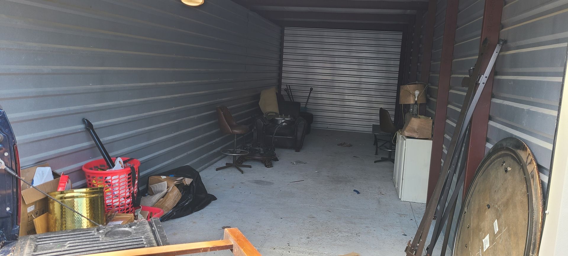 Storage unit with scattered tools, a red bucket, and furniture inside a cluttered metal garage.