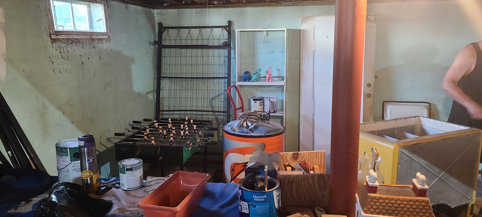 Cluttered room with shelves, cookware, orange bin, and a person standing near a table