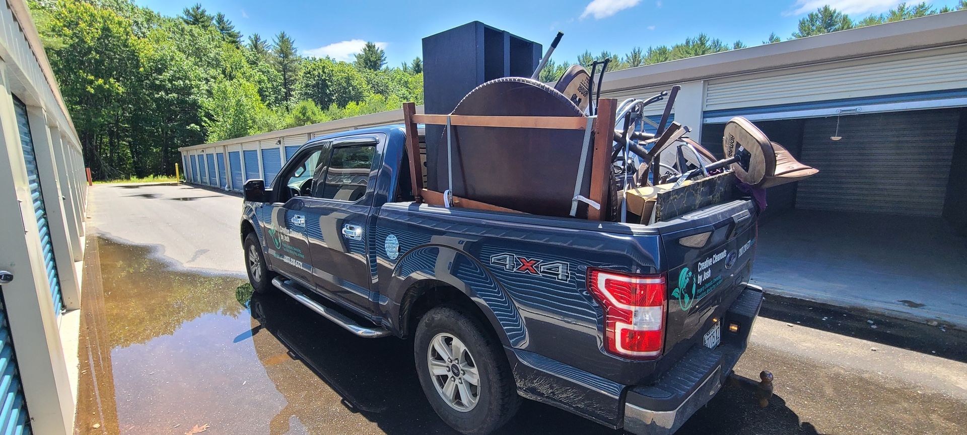 Dark blue pickup truck loaded with furniture and items, parked beside a building in a sunlit driveway.