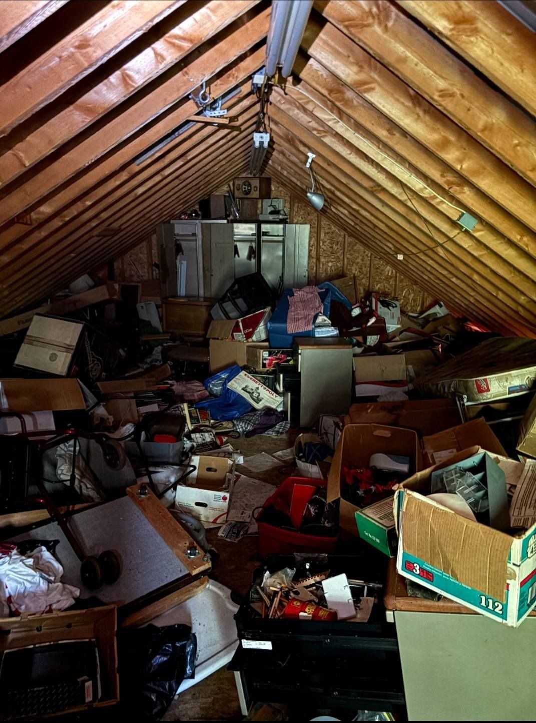 Attic cluttered with boxes, papers, furniture, and scattered debris under slanted wooden rafters.