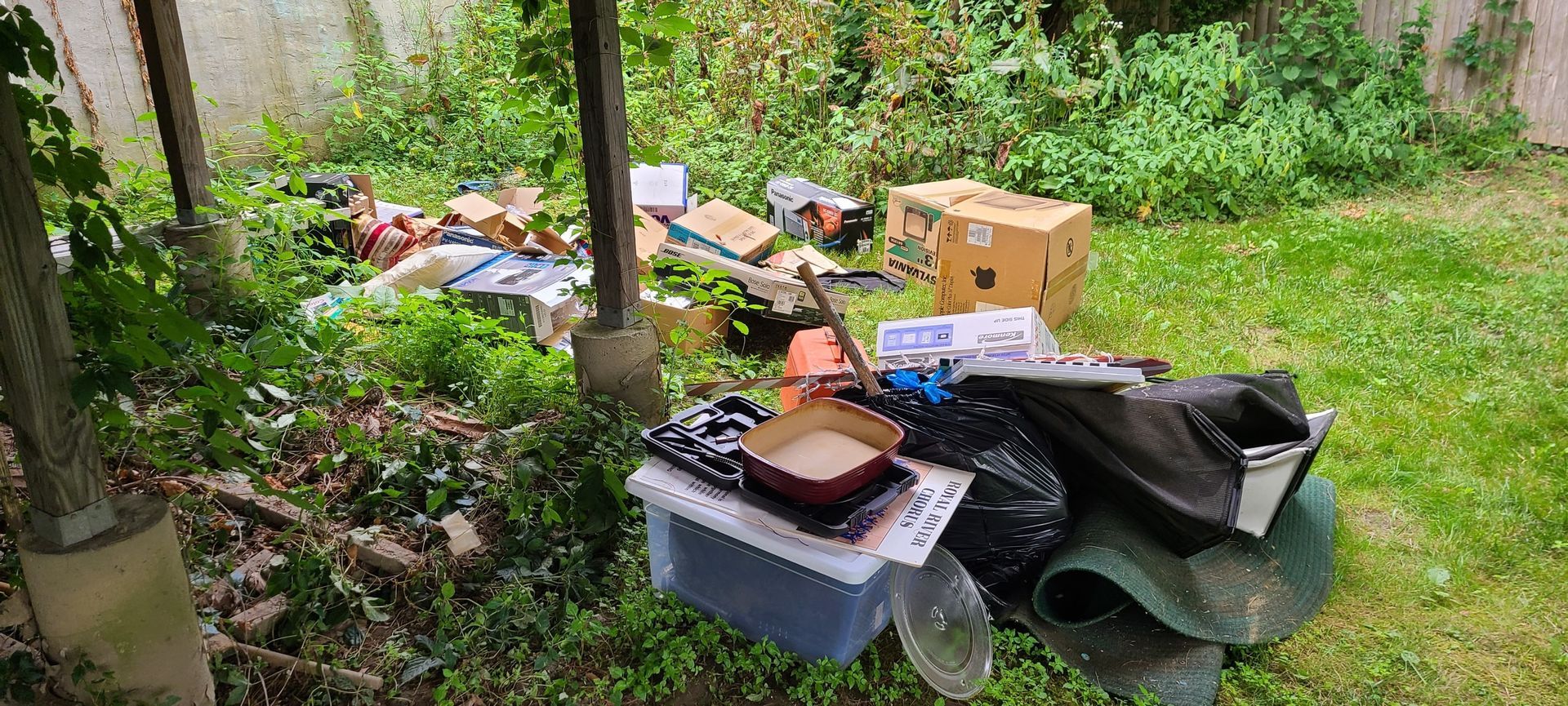Overflowing trash and debris scattered in a grassy outdoor area with bins and cardboard boxes