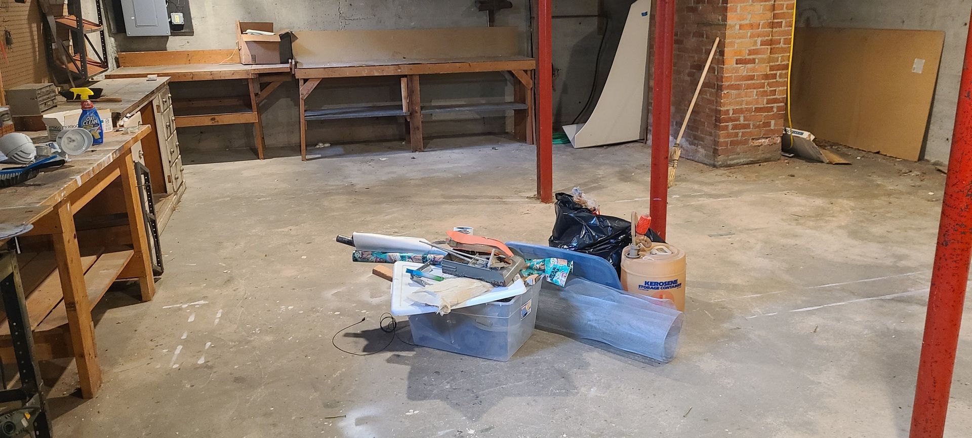 Cluttered workshop with tools and storage bins on a concrete floor, red support posts, and workbenches along the walls
