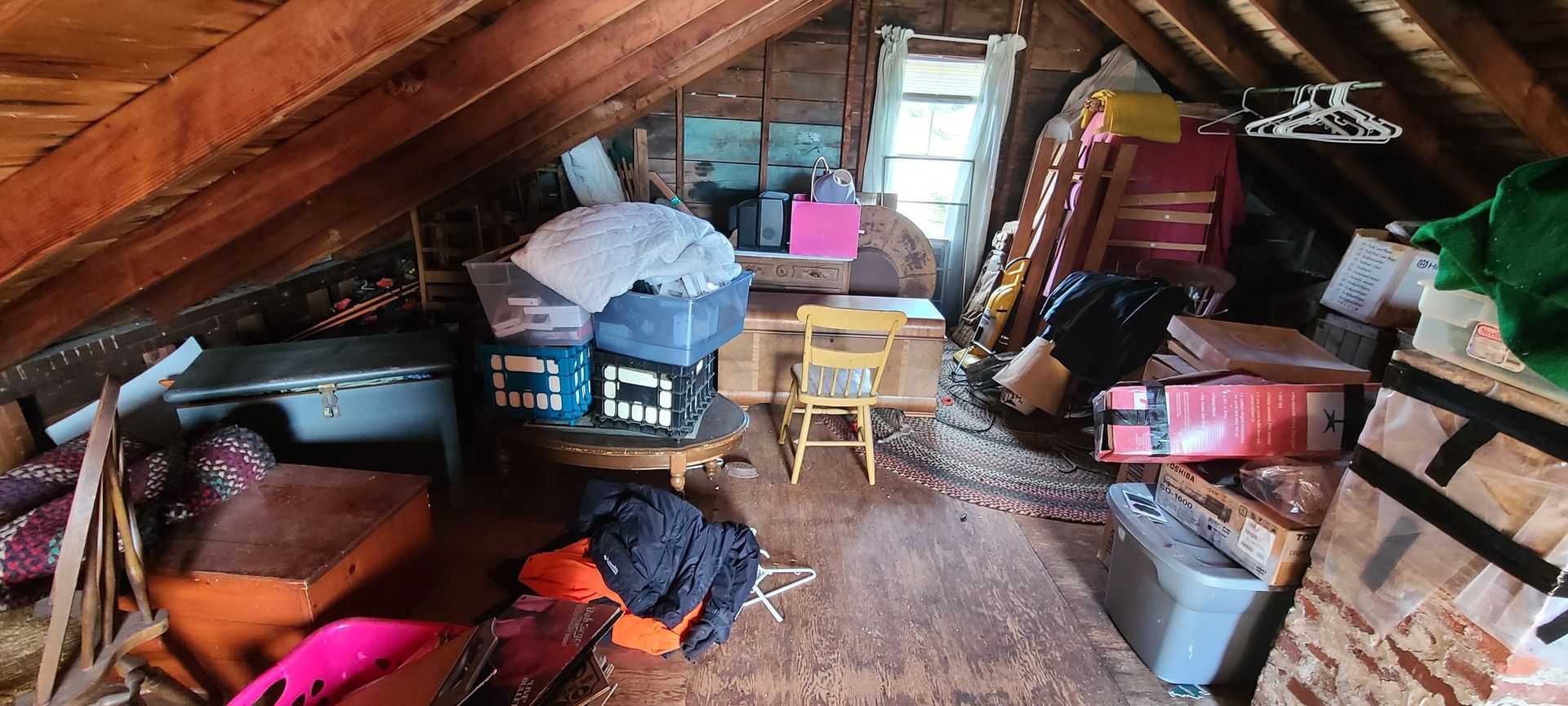Cluttered attic with storage bins, boxes, chairs, tools, and clothes under a sloped wooden roof