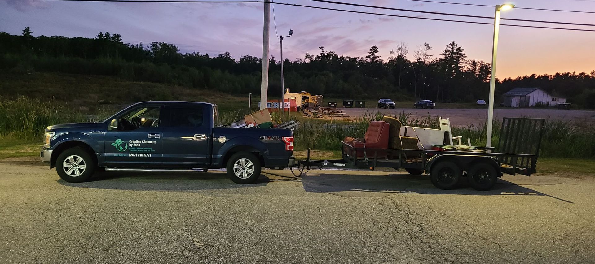 Blue pickup truck towing a flatbed trailer at dusk in a gravel lot, with a fence and streetlights behind it
