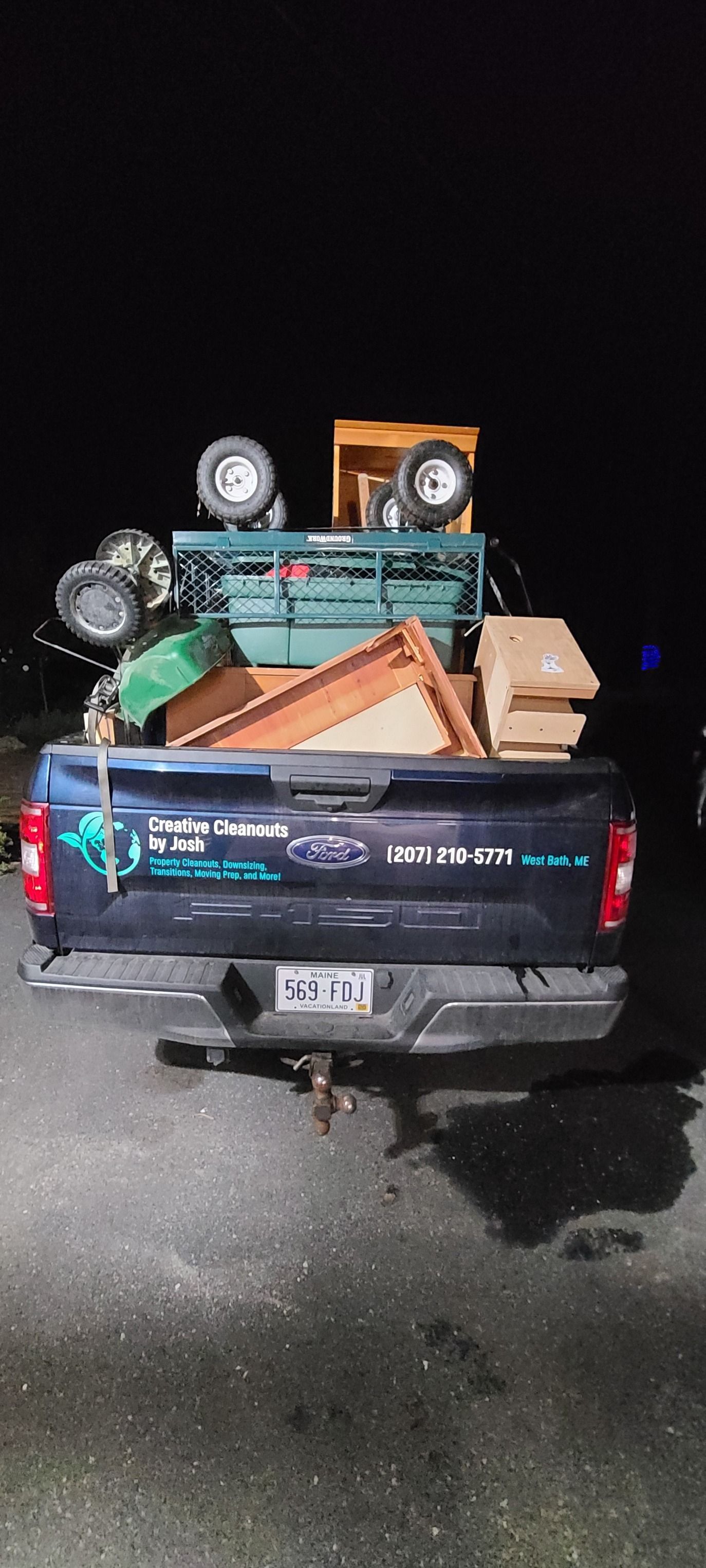 Car trunk open at night, packed with tools, boxes, and other cargo on a wet road.