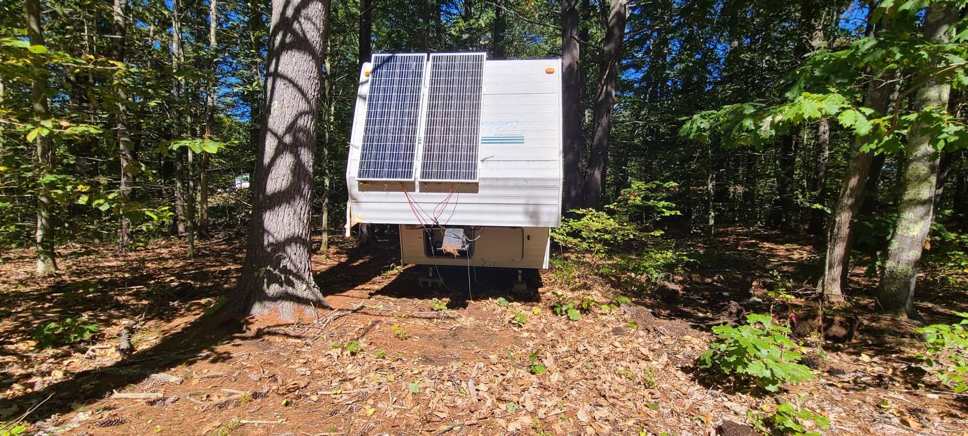 Small white trailer with solar panels in a wooded forest clearing, surrounded by trees and leaves.