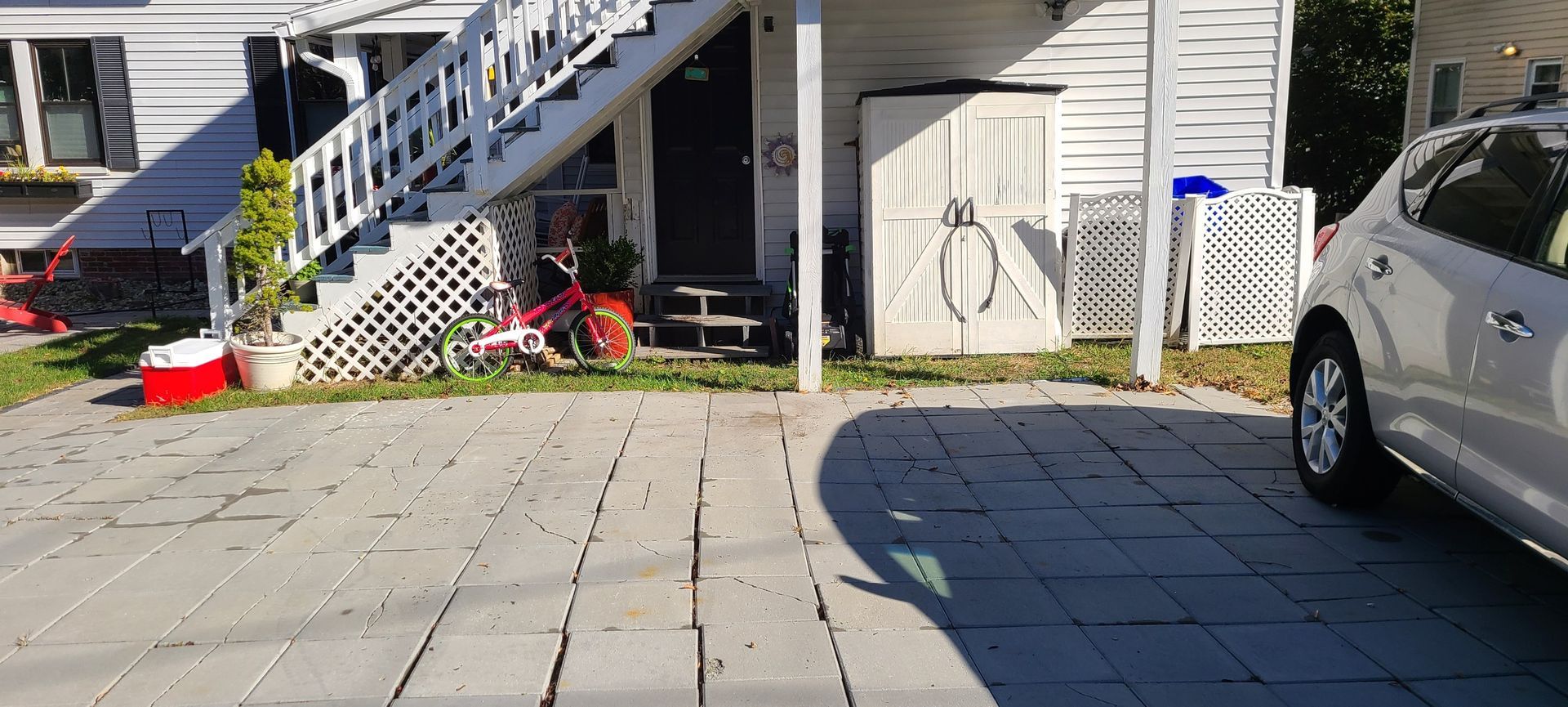Sunny driveway with parked car, porch steps, bike, and outdoor items beside a house