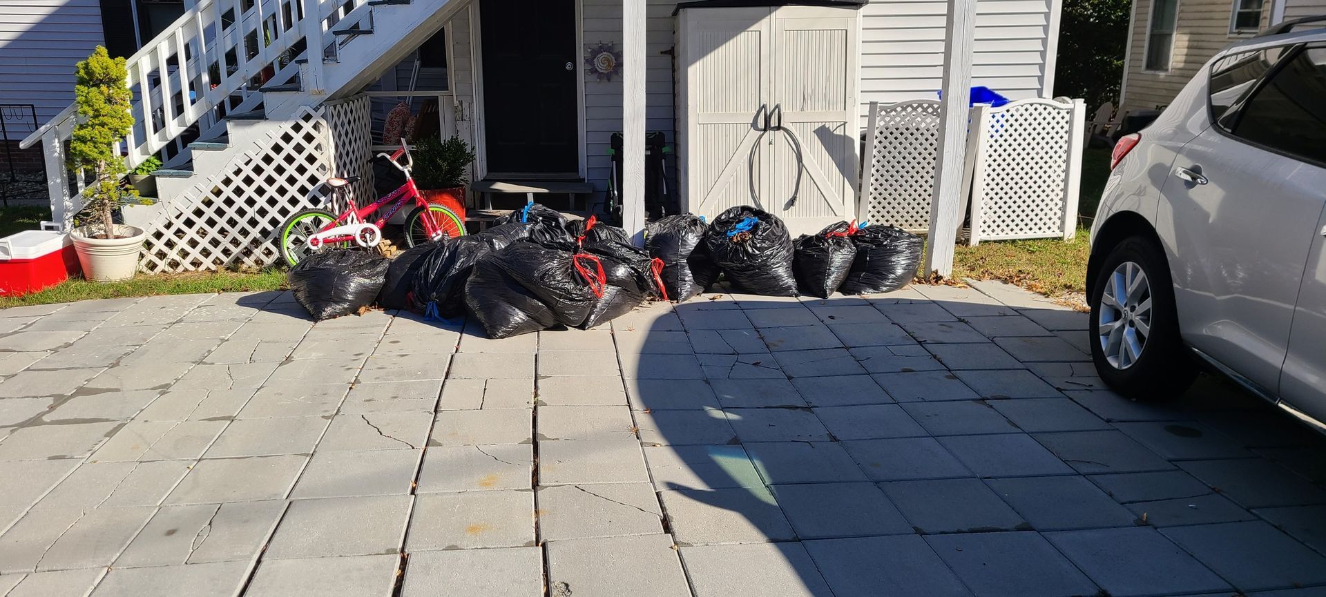 Trash bags piled beside a white building and parked cars in a sunny driveway.