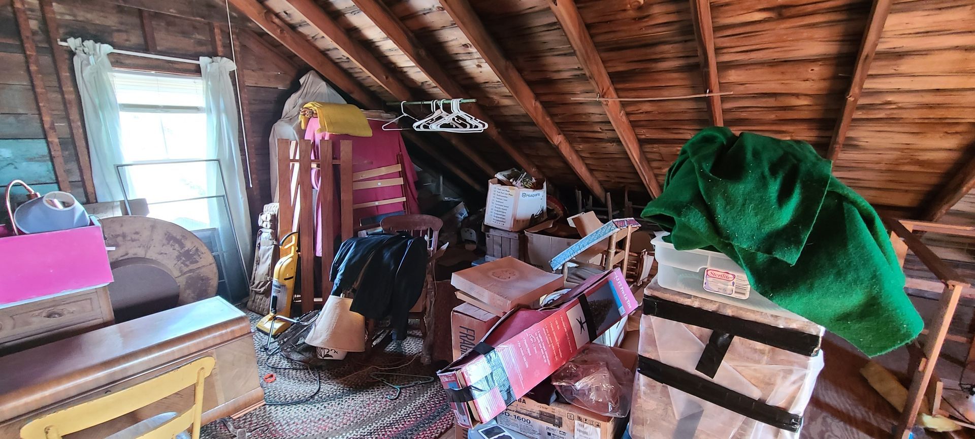 Cluttered attic with clothes, boxes, and furniture under a sloped wooden roof, lit by a bright window