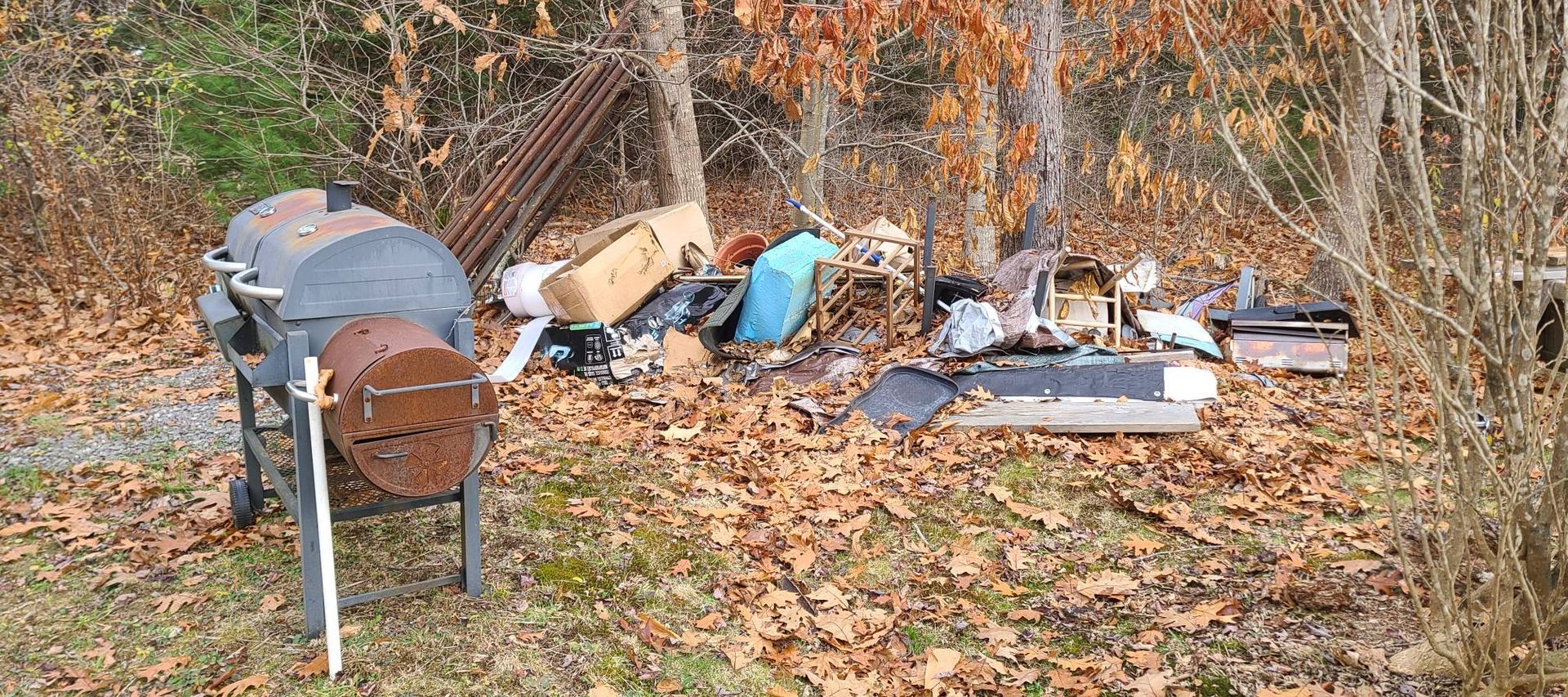 Pile of discarded items and debris along a leaf-covered roadside in a wooded area, with a barrel in the foreground.